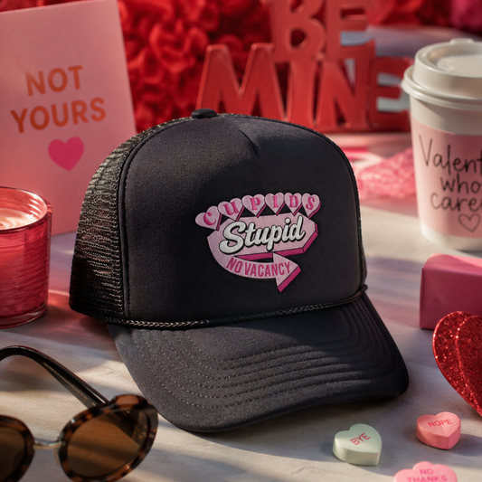 A black trucker hat with a pink and white embroidered graphic reading 'Cupid is Stupid' surrounded by Valentine's Day themed props including heart candy, sunglasses, a coffee cup, and a sign that says 'NOT YOURS'.