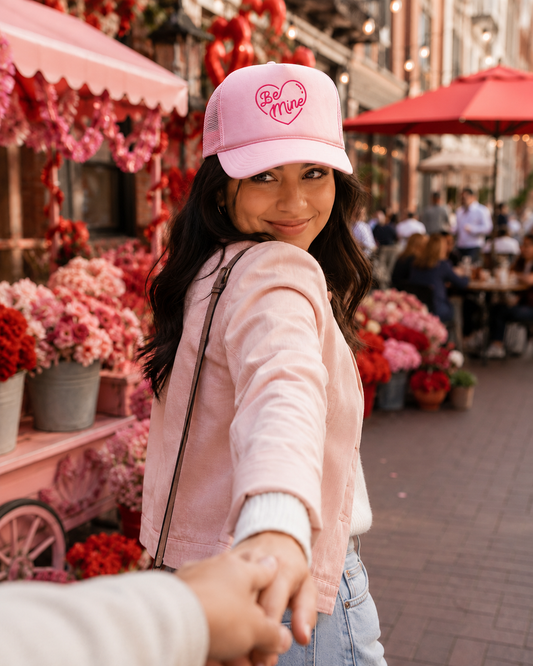 A woman in a pink 'Be Mine' trucker hat and a light pink jacket holds hands with someone in the foreground, set in a vibrant outdoor flower market scene.