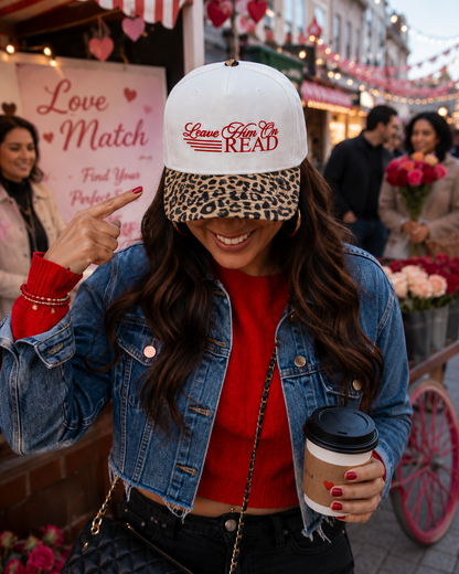 A woman wearing an off-white baseball cap with a leopard-print brim and red embroidered text reading 'Leave Him on Read' points to the hat while holding a coffee cup in an outdoor market setting.
