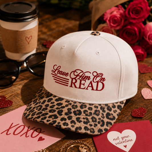 An off-white snapback cap with a leopard-print brim and red embroidered text reading 'Leave Him On READ,' styled on a wooden table with a coffee cup, sunglasses, red roses, and Valentine-themed cards.