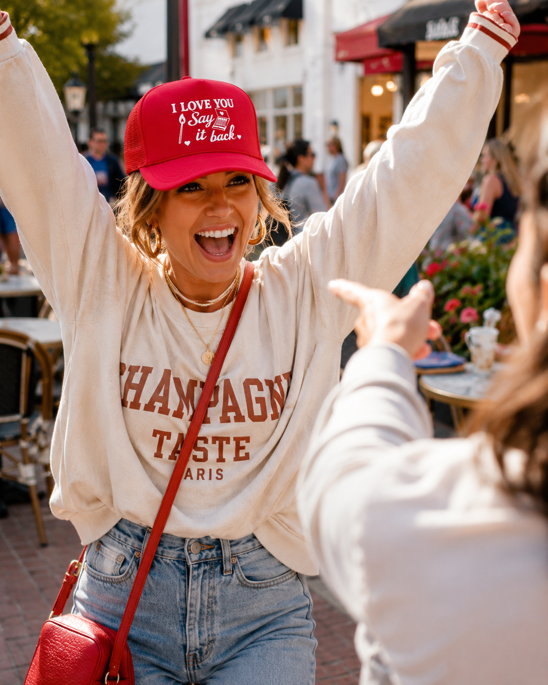 A woman outdoors wearing a cream sweatshirt with text in brown, a red baseball cap with white text, and light blue jeans, smiling with raised arms while another person points toward her.