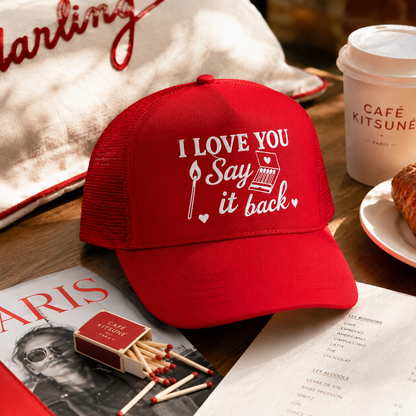 A red trucker-style baseball cap with white embroidered text reading 'I LOVE YOU Say it back' and a small typewriter graphic, styled on a wooden table with a coffee cup, magazine, matches, and a plate in a cafe setting.
