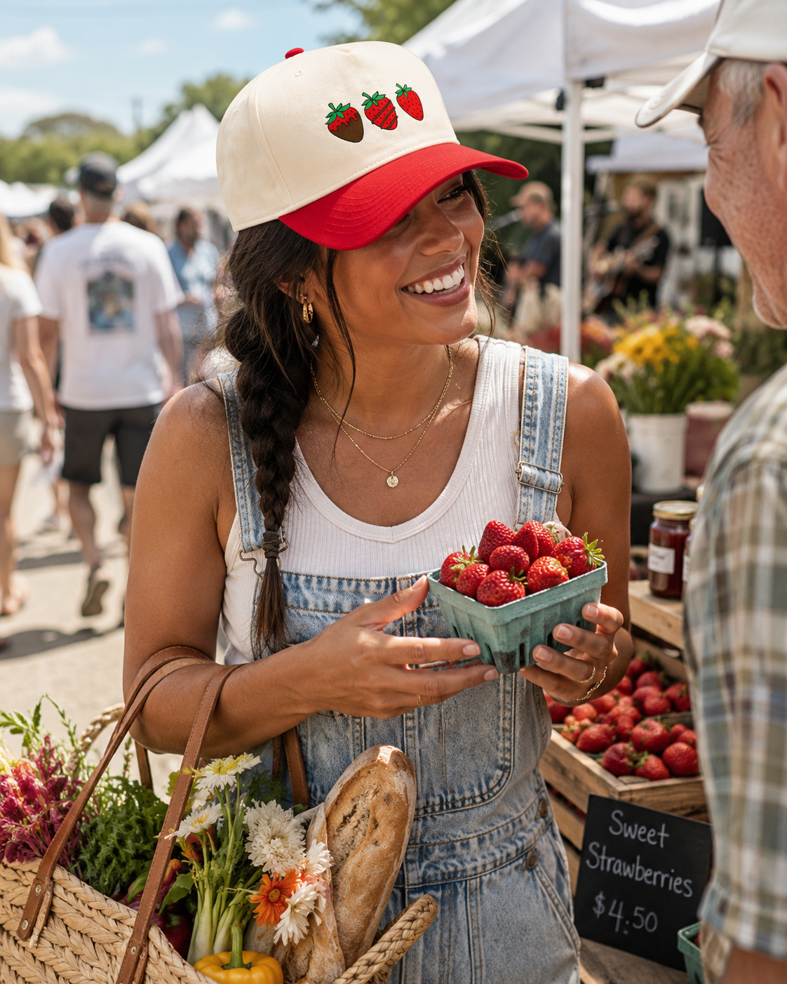 A woman at an outdoor farmers market wears a cream and red trucker hat with embroidered strawberries, denim overalls, and a white tank top while holding a basket of fresh strawberries.