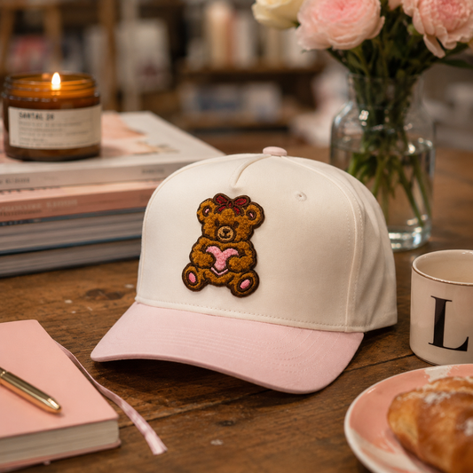 A off-white baseball cap with a light pink brim, featuring a chenille, brown teddy bear patch with a pink heart on the front. The cap is styled on a wooden table with a pink notebook, a lit candle, a vase of pink roses, and a pastry in the background.