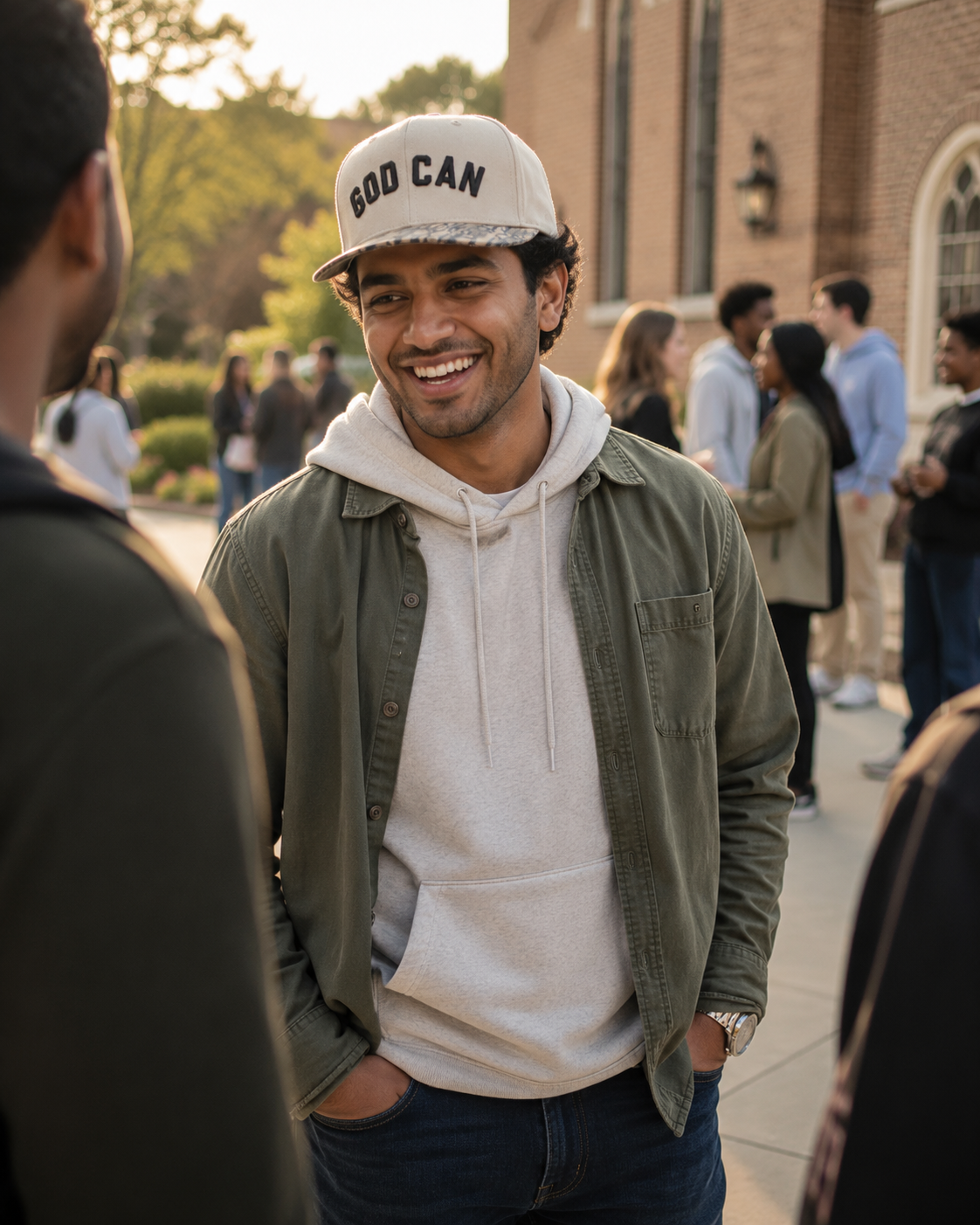 A smiling man outdoors wearing a beige baseball cap with 'GOD CAN' printed on the front, an olive green overshirt, and a light beige hoodie. The background shows a brick building and other people in a campus-like setting.