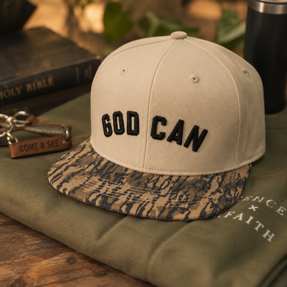 A beige snapback cap with black embroidered 'GOD CAN' text on the front and a camouflage-patterned visor, styled on an olive green sweatshirt with a Bible and keychain in the background.