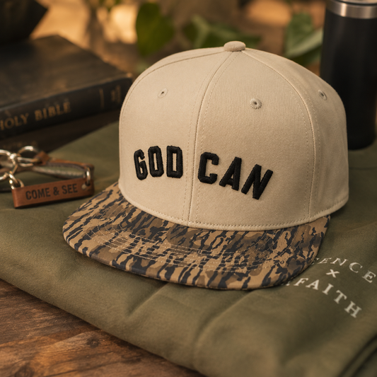 A beige snapback cap with black embroidered 'GOD CAN' text on the front and a camouflage-patterned visor, styled on an olive green sweatshirt with a Bible and keychain in the background.