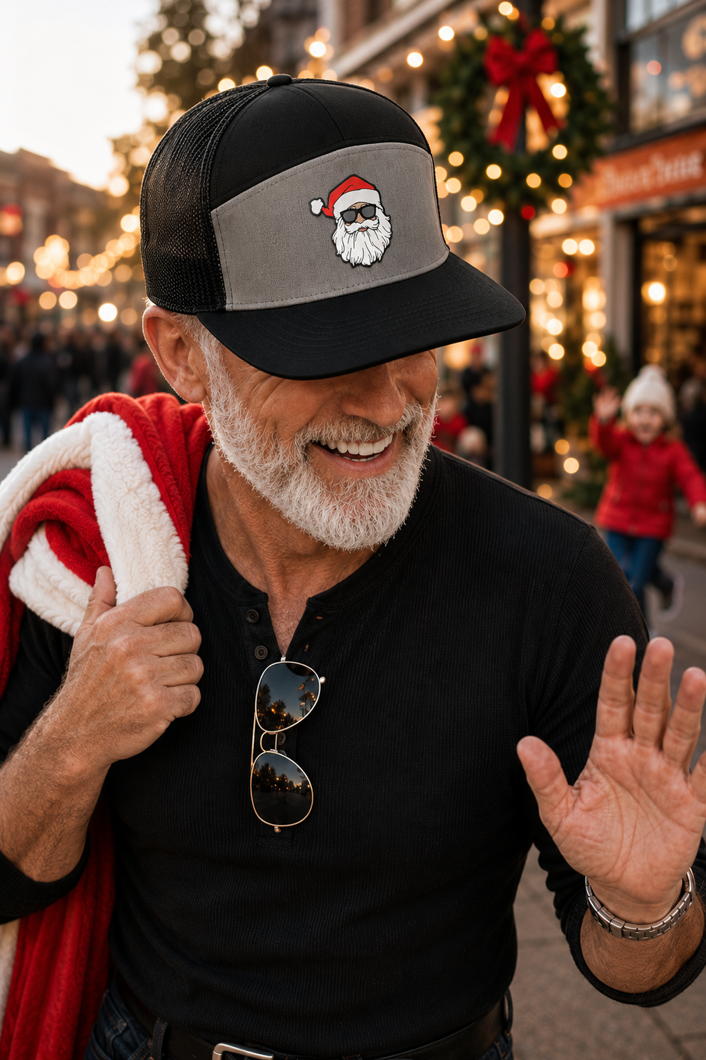 A smiling man wearing a black and gray trucker hat with a Santa face graphic on the front panel, standing outdoors in a festive holiday street scene with lights and garlands in the background.