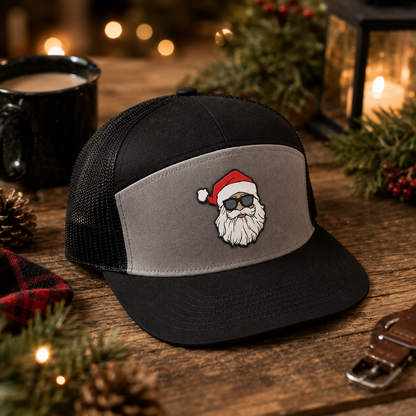 A black and gray trucker hat with a custom embroidered Santa Claus face on the front panel, displayed on a rustic wooden table with holiday decorations, a mug, and a lantern in the background.