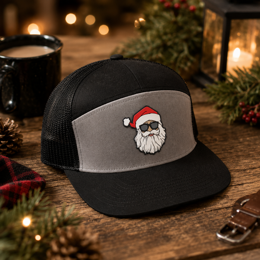 A black and gray trucker hat with a custom embroidered Santa Claus face on the front panel, displayed on a rustic wooden table with holiday decorations, a mug, and a lantern in the background.