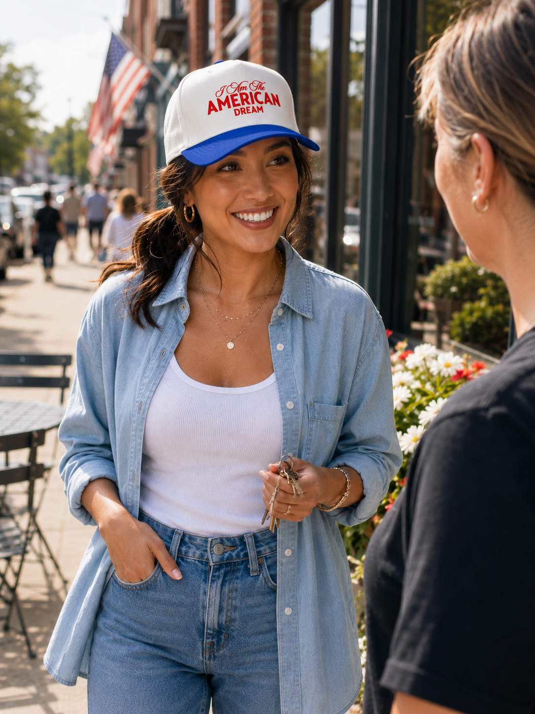 A woman outdoors wearing a white and blue baseball cap with 'I am the AMERICAN DREAM' printed on the front, a light blue button-up shirt, and a white tank top, smiling and talking to another person.