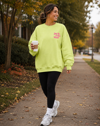 A woman walking on an outdoor path wearing an oversized neon yellow-green sweatshirt with pink text on the chest, black leggings, and white sneakers, while holding a coffee cup.