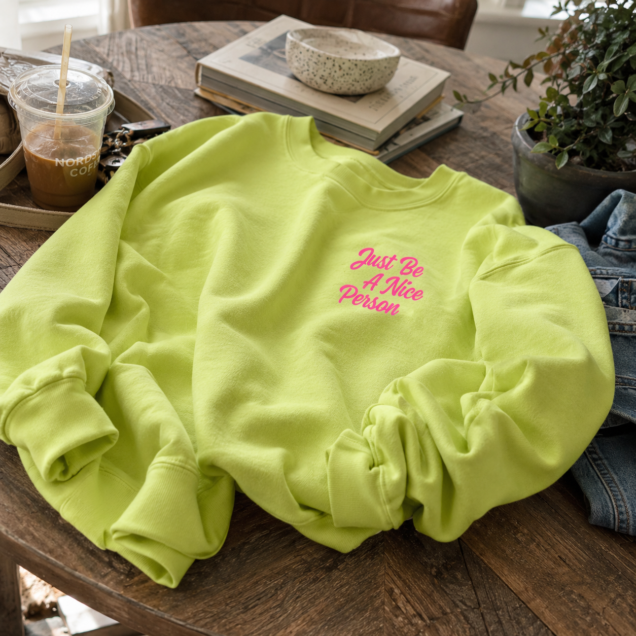 A bright lime green crewneck sweatshirt with pink embroidered text reading 'Just Be A Nice Person' on the left chest, styled on a rustic wooden table with books, a plant, and a denim jacket nearby.
