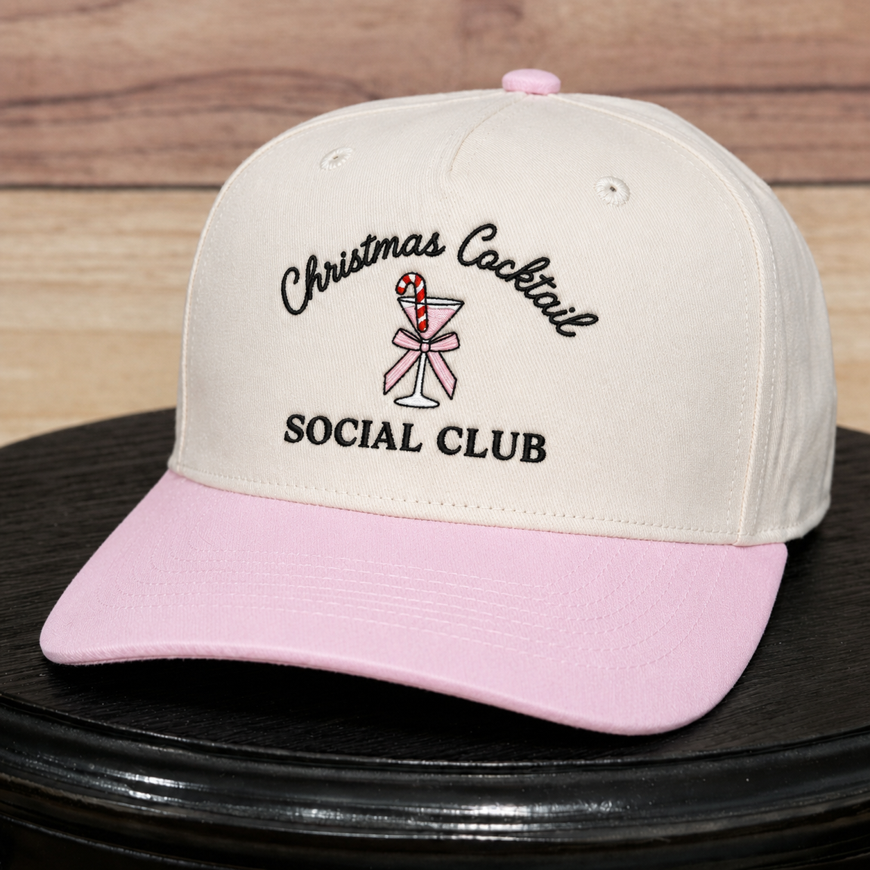 A cream-colored baseball cap with a light pink brim, featuring embroidered text that reads 'Christmas Cocktail SOCIAL CLUB' with a pink and white martini glass graphic, displayed on a black surface against a wooden background.