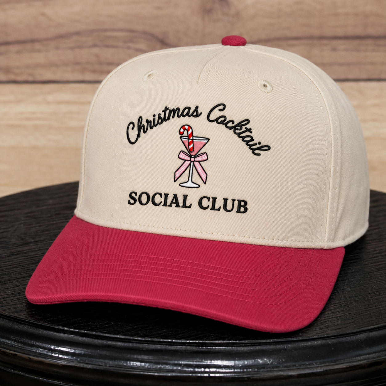 A cream-colored baseball cap with a bright pink brim, featuring embroidered text that reads 'Christmas Cocktail SOCIAL CLUB' around a pink bow and martini glass graphic. The cap is displayed on a black surface with a rustic wood background.
