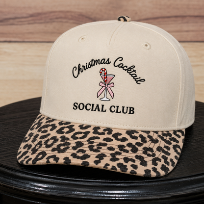A beige baseball cap with a leopard-print brim, featuring embroidered text that reads 'Christmas Cocktail SOCIAL CLUB' with a cocktail glass and candy cane graphic on the front.