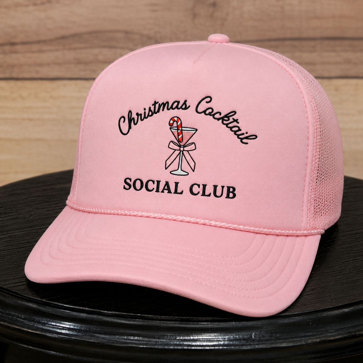 A light pink trucker-style cap with embroidered text reading 'Christmas Cocktail SOCIAL CLUB' and a cocktail graphic, displayed on a black surface against a wood plank background.