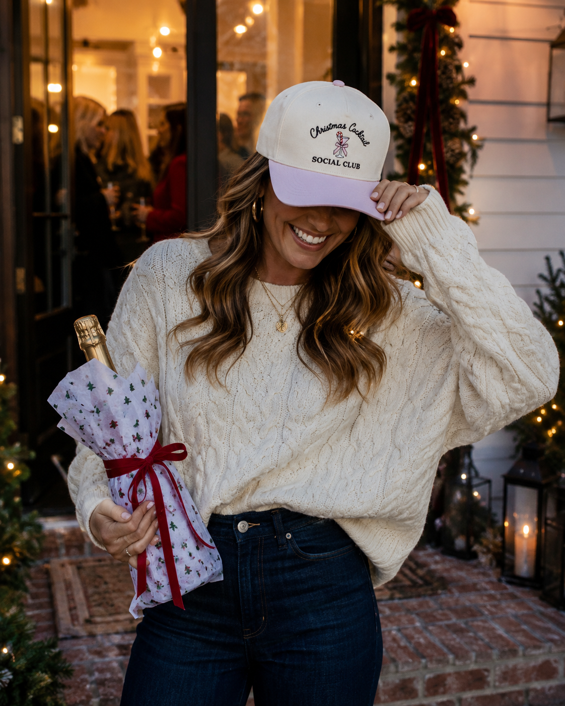 A woman wearing a cream cable-knit sweater, dark blue jeans, and a cream and lavender baseball cap with 'Christmas Cocktail Social Club' embroidered on it. She is holding a wrapped champagne bottle in a festive, warmly lit storefront setting.
