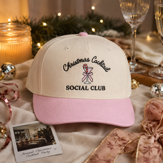 A cream and light pink baseball cap with embroidered text reading 'Christmas Cocktail SOCIAL CLUB' and a martini glass with a candy cane, styled on a festive table with candles, glasses, ribbons, and holiday decor.