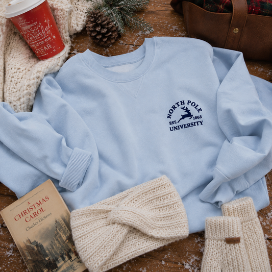 A light blue crewneck sweatshirt with a navy 'North Pole University' logo, styled in a cozy flat-lay arrangement with a red Christmas mug, knit accessories, a book, and pinecones on a wooden surface.