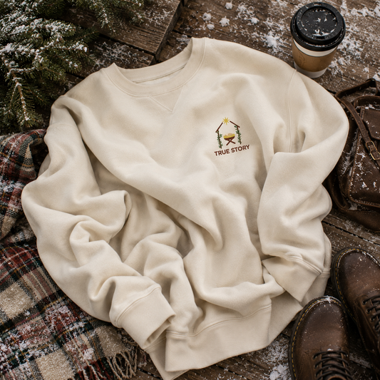 An off-white crewneck sweatshirt with a small embroidered campfire and the words 'TRUE STORY' on the chest, styled in a cozy flat-lay with a coffee cup, boots, and a plaid blanket.