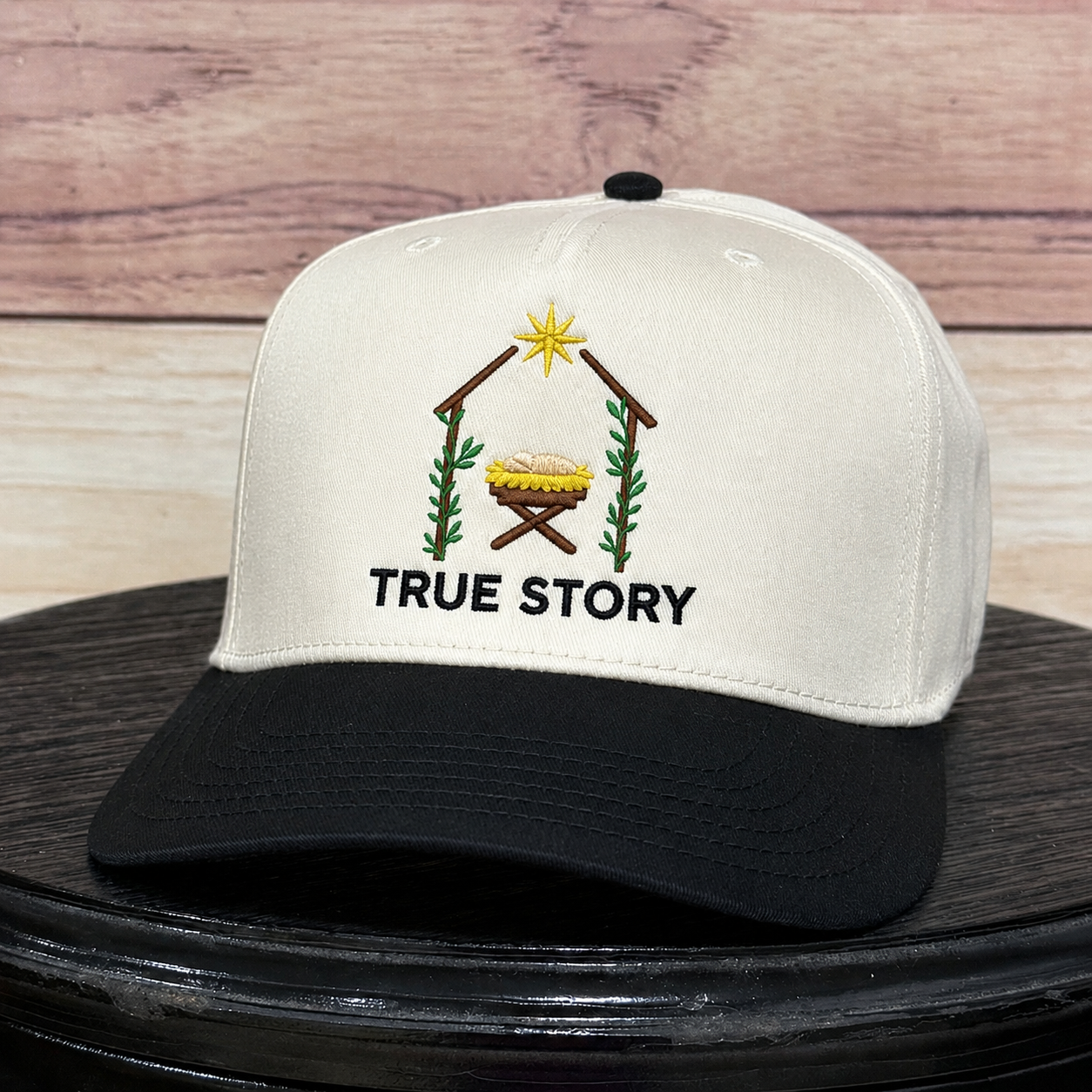 An off-white and black baseball cap with an embroidered nativity scene and the text 'TRUE STORY' on the front, displayed on a black surface against a rustic wood plank background.