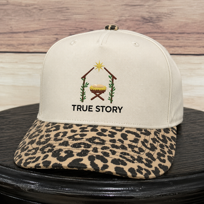 A structured baseball cap with an off-white crown and a leopard-print visor, featuring an embroidered nativity scene and the text 'TRUE STORY' on the front. The cap is displayed on a black surface with a rustic wood background.