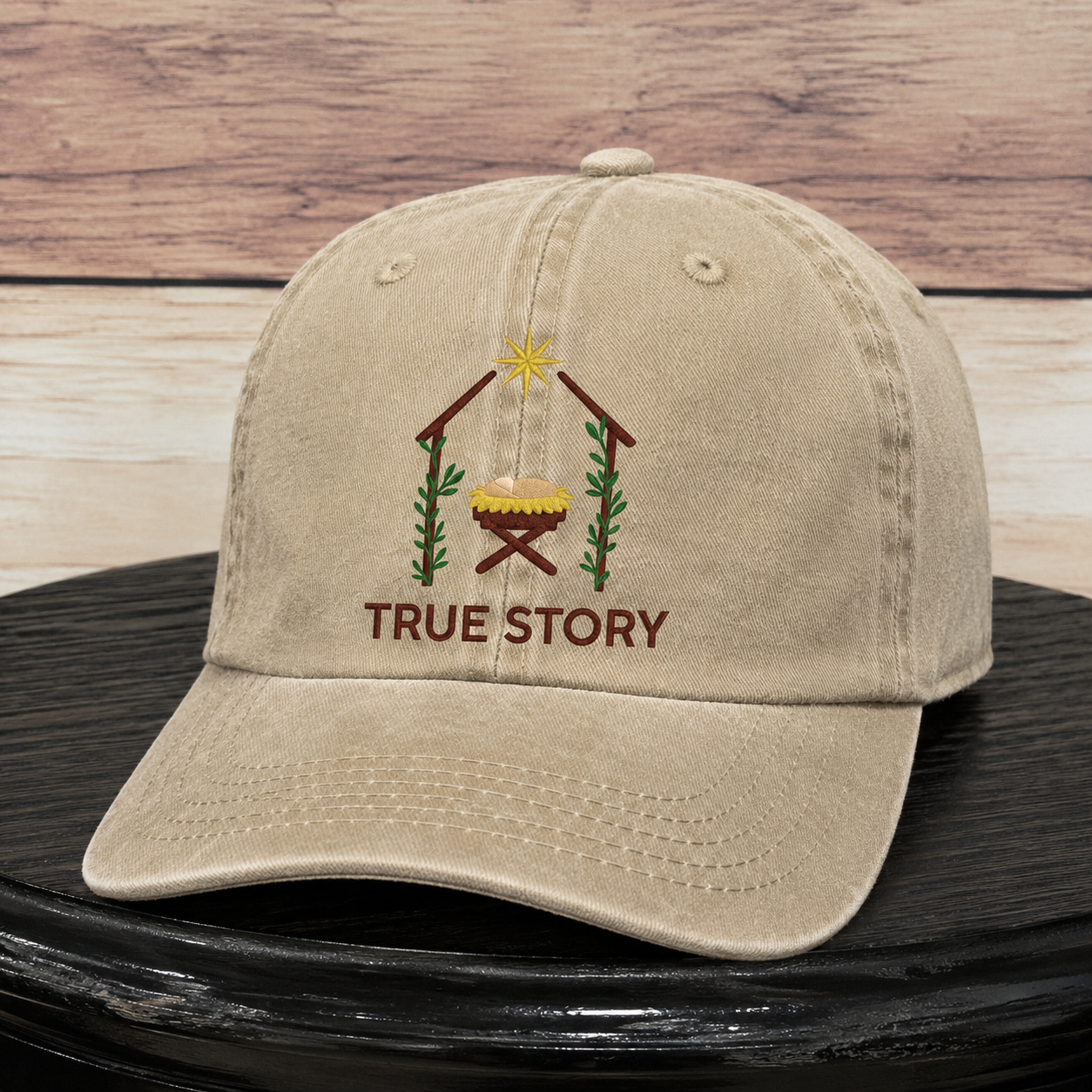 A beige cotton baseball cap with an embroidered Christmas nativity scene and the words 'TRUE STORY' on the front, displayed on a black surface against a rustic wood plank background.