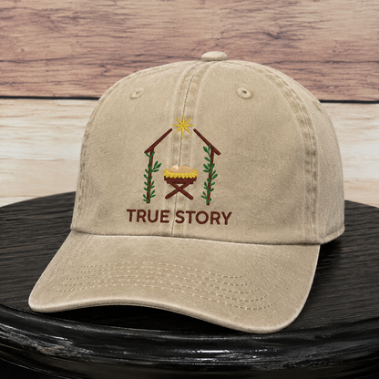 A beige cotton baseball cap with an embroidered Christmas nativity scene and the words 'TRUE STORY' on the front, displayed on a black surface against a rustic wood plank background.