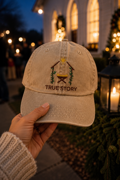 A hand holds a beige distressed baseball cap featuring an embroidered nativity scene and the text 'TRUE STORY' against a blurred outdoor holiday background with warm lights.