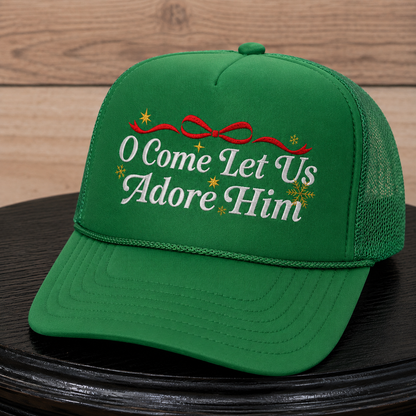 A green trucker-style baseball cap with embroidered white text reading 'O Come Let Us Adore Him,' a red bow, and small yellow star and snowflake motifs, displayed on a dark round surface against a wood plank background.