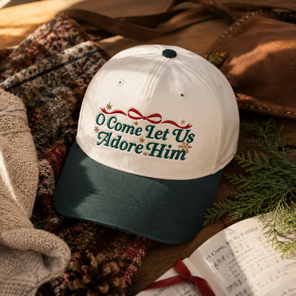 A baseball cap with a white crown and dark green brim, embroidered with the phrase 'O Come Let Us Adore Him' in green script with a red bow and small gold stars. The cap is styled in a cozy flat-lay arrangement with a knit sweater, brown leather, pine branches, and an open book.