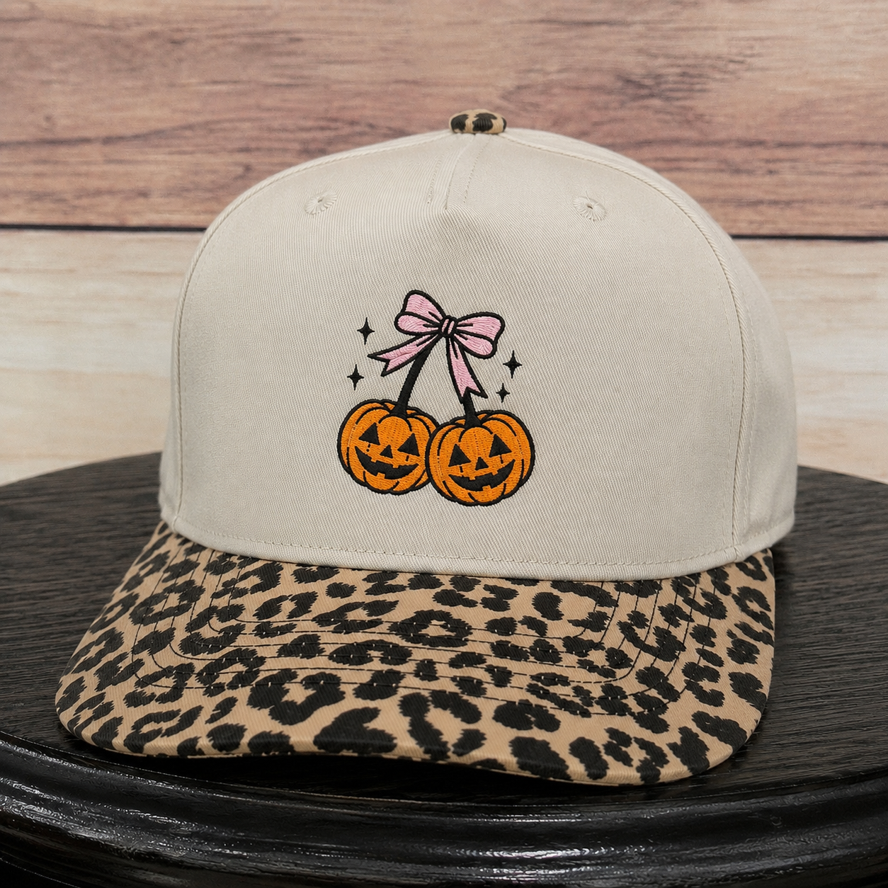 A beige snapback cap with a leopard-print flat brim, featuring an embroidered design of two jack-o'-lanterns tied with a pink bow on the front.