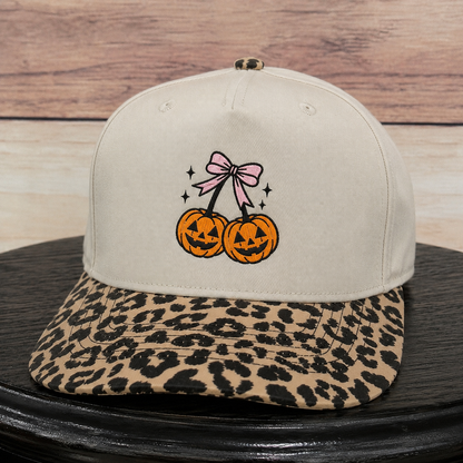 A beige snapback cap with a leopard-print flat brim, featuring an embroidered design of two jack-o'-lanterns tied with a pink bow on the front.