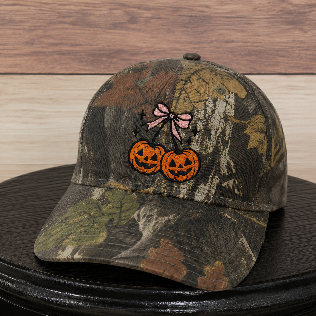 A camouflage baseball cap with an embroidered design of two orange jack-o'-lantern pumpkins tied together by a pink bow, displayed on a black round surface against a wood-paneled background.