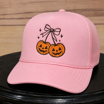A light pink trucker cap with an embroidered design of two jack-o'-lanterns hanging from a bow, displayed on a black surface against a wood background.