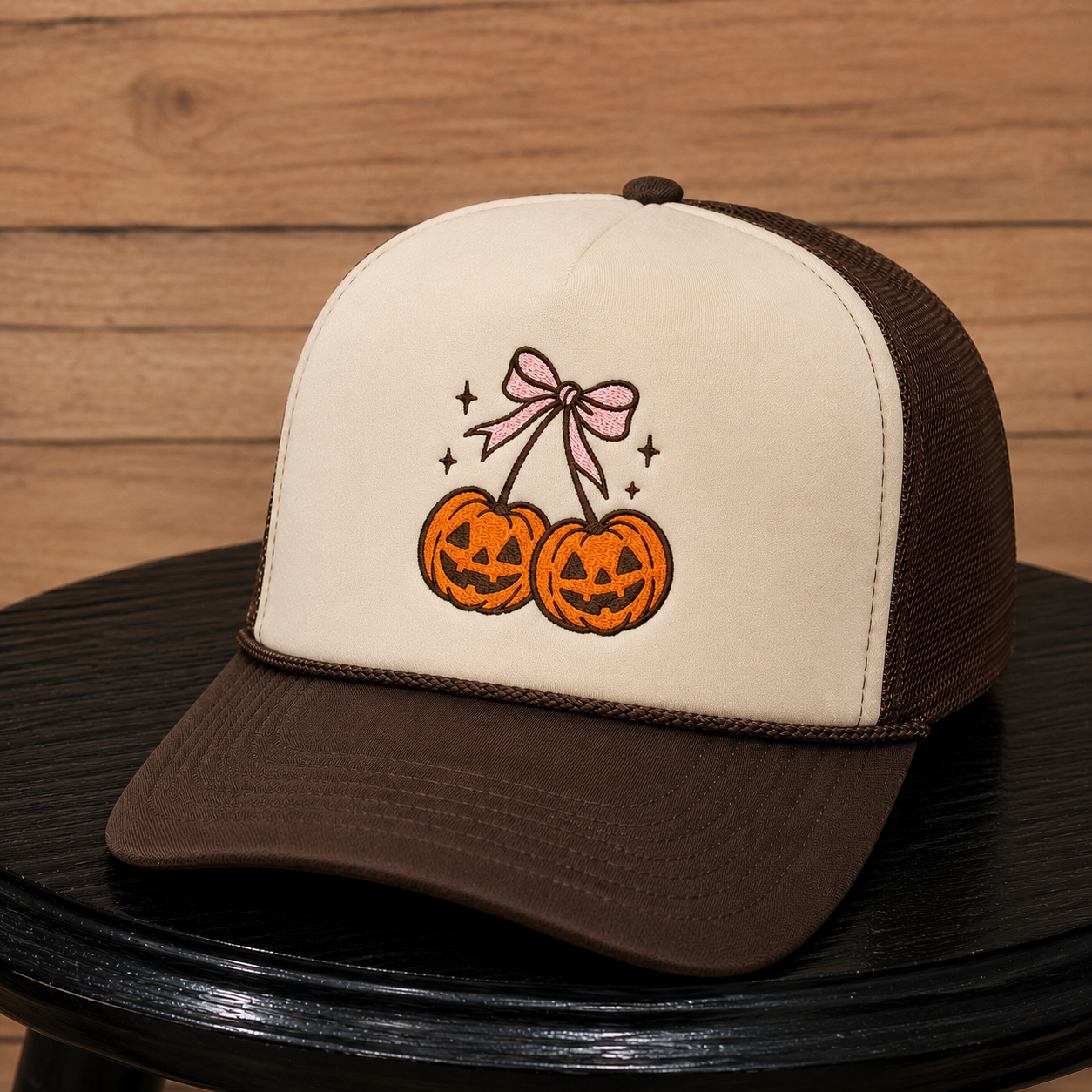 A brown and cream trucker hat with an embroidered design of two jack-o'-lanterns on a pink bow, resting on a black surface against a wooden background.