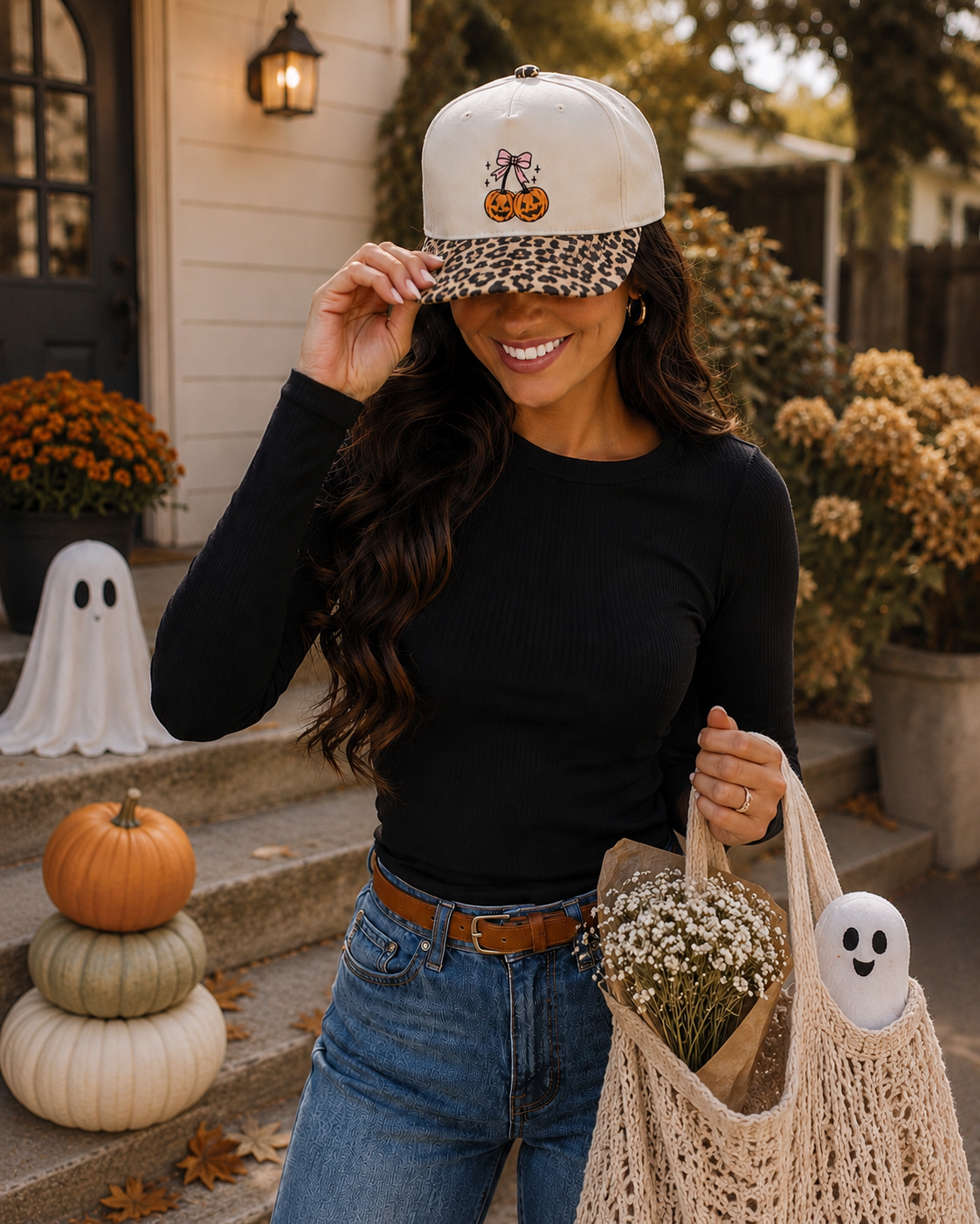 A woman wearing a white baseball cap with a leopard-print brim and a small jack-o'-lantern graphic, posing on outdoor steps with autumn decorations, a ghost bag, and a black long-sleeve top.