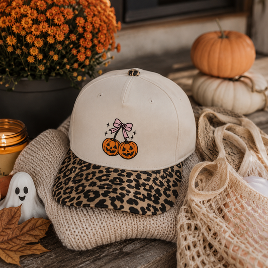 An off-white baseball cap with a leopard-print visor and embroidered orange pumpkins under a pink bow, styled on a knit blanket with autumn props including pumpkins, flowers, a small ghost, and a net bag.
