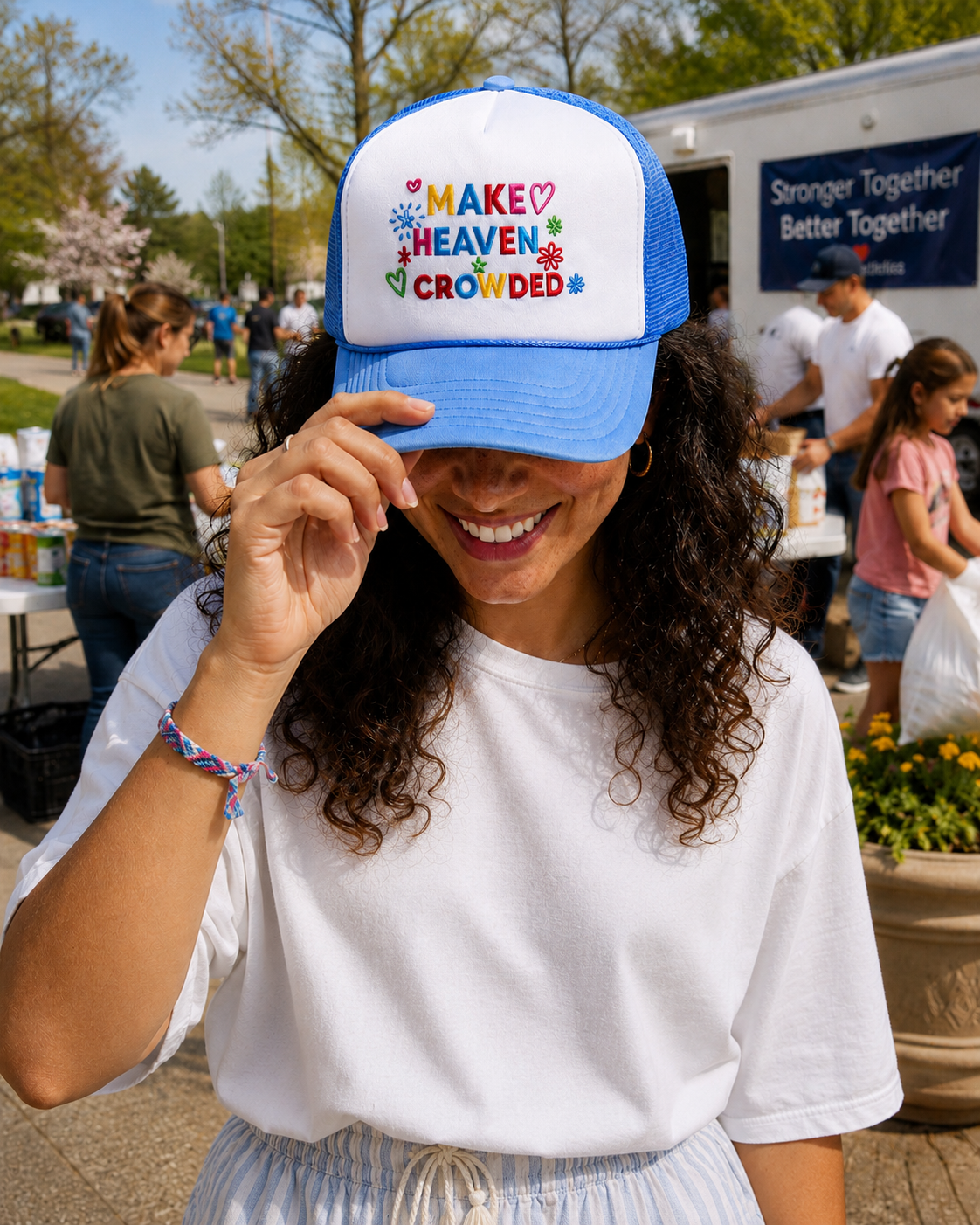 A woman outdoors wearing a blue and white trucker hat with the text 'MAKE HEAVEN CROWDED' in colorful letters. The hat has blue front panels and white mesh back panels.