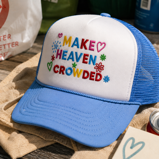 A blue and white trucker hat with colorful embroidered text reading 'MAKE HEAVEN CROWDED' and small heart and flower motifs, styled on a burlap surface with other casual props in the background.