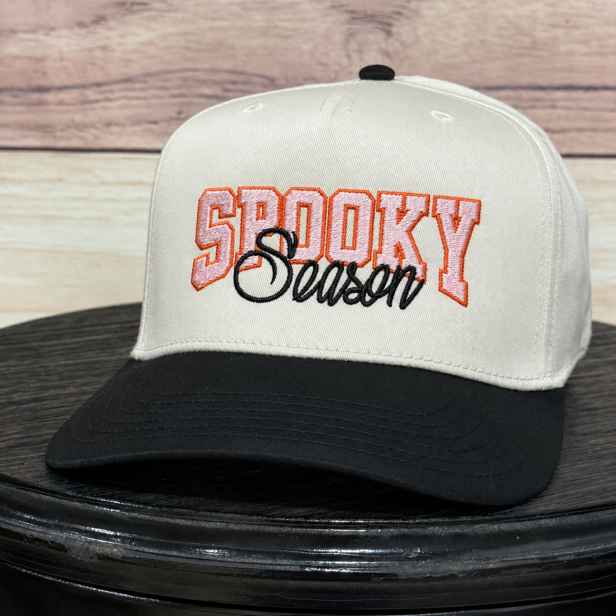 A baseball cap with an off-white crown and black brim, featuring 'SPOOKY Season' embroidered on the front. The cap is displayed on a black round surface with a light wood background.