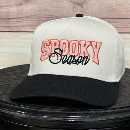 A baseball cap with an off-white crown and black brim, featuring 'SPOOKY Season' embroidered on the front. The cap is displayed on a black round surface with a light wood background.