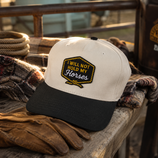 A baseball cap with an off-white crown and black brim, featuring a decorative embroidered patch that reads 'I WILL NOT HOLD MY Horses' in yellow and black. The cap is styled on a rustic wooden surface with leather work gloves and a plaid blanket in the background.