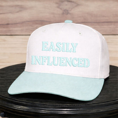 Beige and light blue cap with 'EASILY INFLUENCED' text on a wooden surface