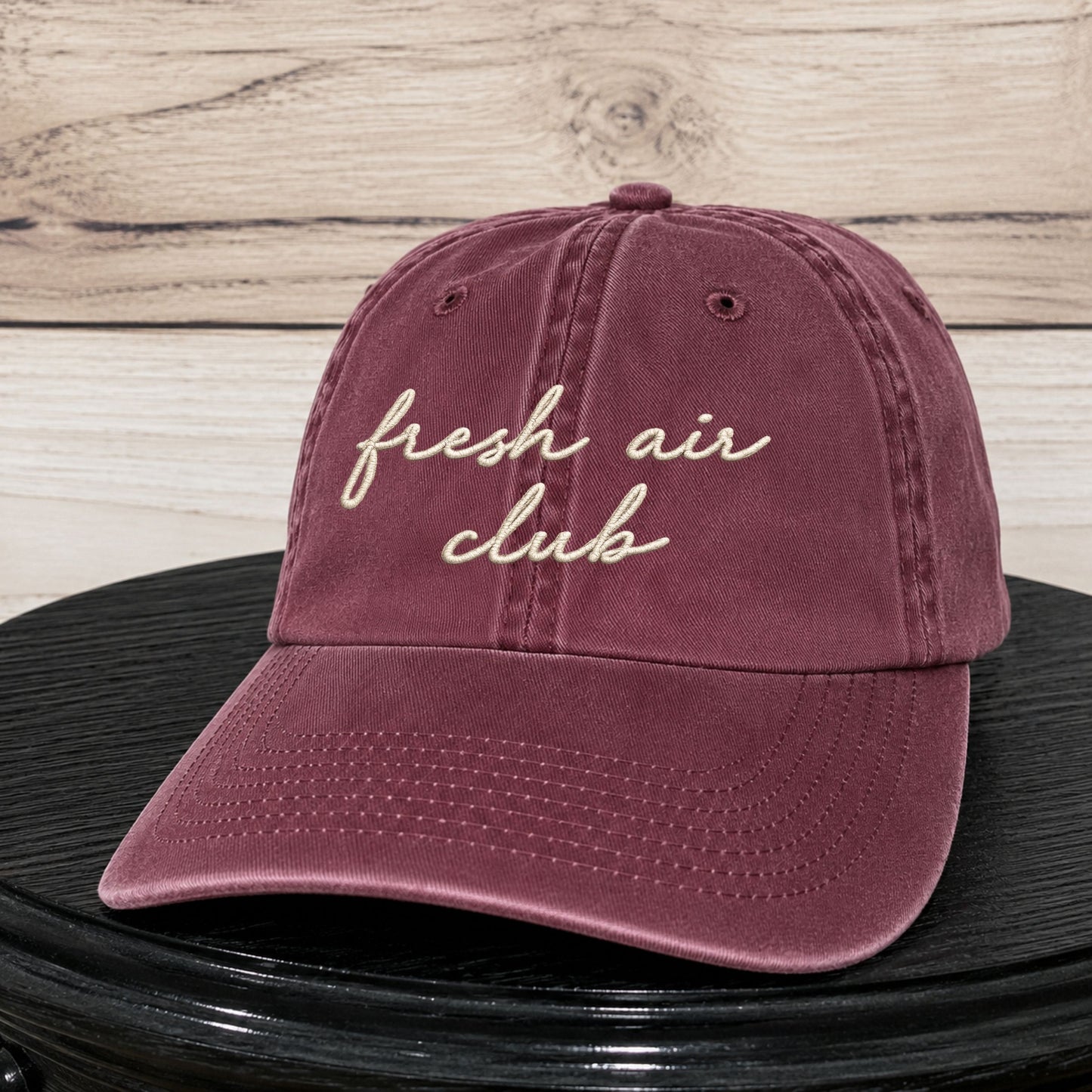 A dusty burgundy baseball cap with 'fresh air club' embroidered in cream script on the front, resting on a black surface against a light wood plank background.