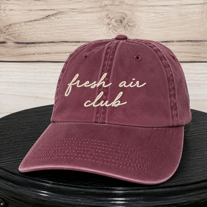 A dusty burgundy baseball cap with 'fresh air club' embroidered in cream script on the front, resting on a black surface against a light wood plank background.