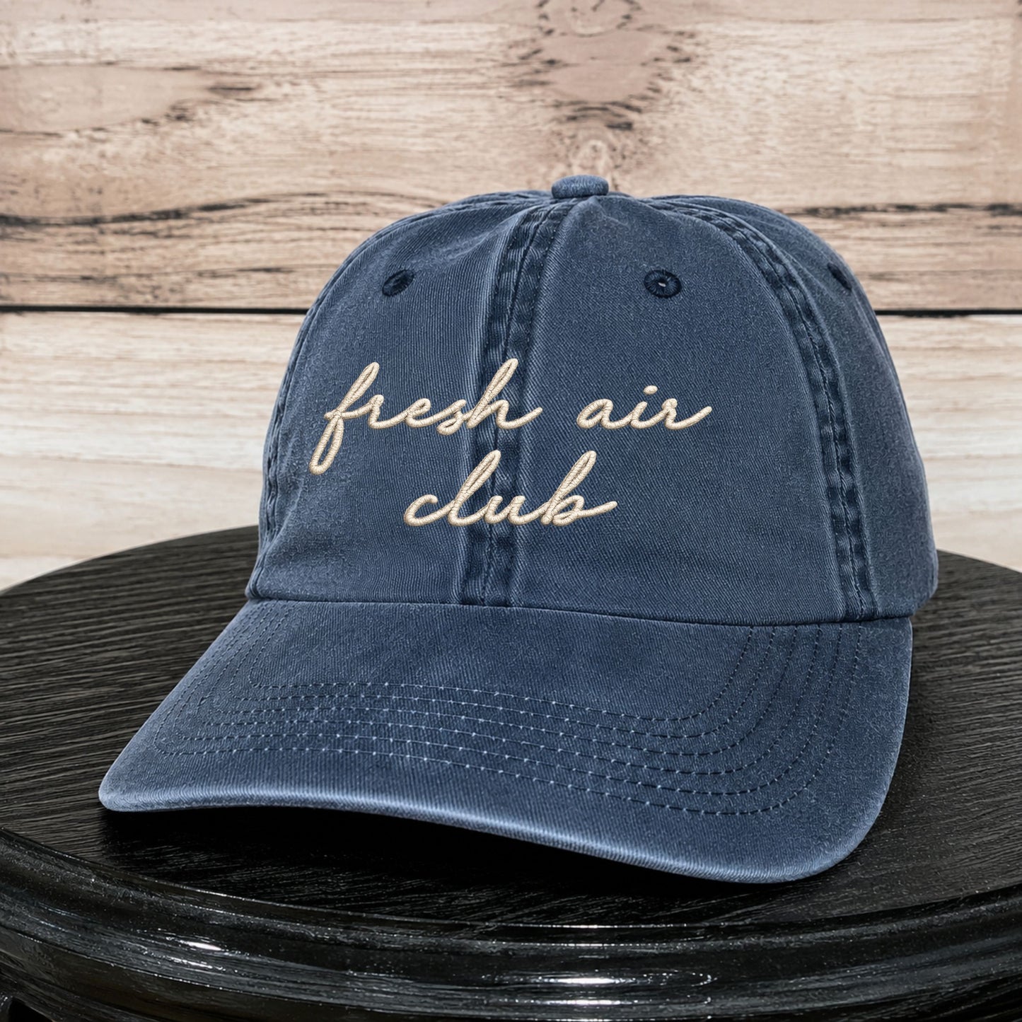 A navy blue washed cotton baseball cap with 'fresh air club' embroidered in beige script across the front, displayed on a black round surface against a light wood plank background.
