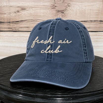 A navy blue washed cotton baseball cap with 'fresh air club' embroidered in beige script across the front, displayed on a black round surface against a light wood plank background.