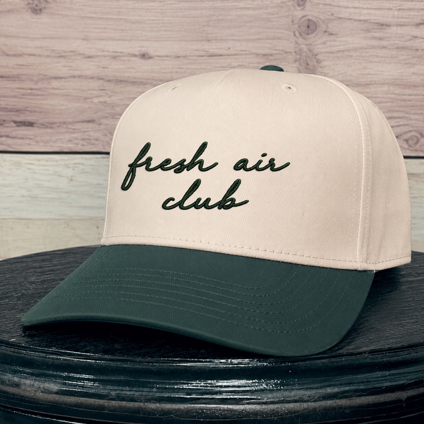A baseball cap with an natural crown and a dark green brim, featuring the text 'fresh air club' embroidered in dark green script on the front. The cap is displayed on a dark wooden surface against a light wood plank background.
